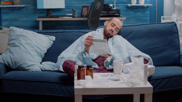 Tired ill person reading medication leaflet to cure cold and flu with prescription treatment. Man with disease feeling unhappy while looking at paper of medicaments against headache