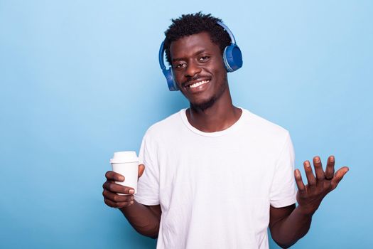 Man wearing headphones while holding cup of coffee and looking at camera. Young adult listening to music with takeaway drink in hand. Person feeling happy enjoying beverage and technology.
