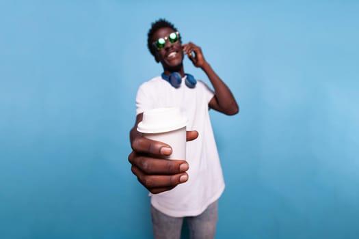 African american man holding cup of coffee while chatting on phone call. Black person with sunglasses and headphones showing drink to camera, talking on smartphone and smiling