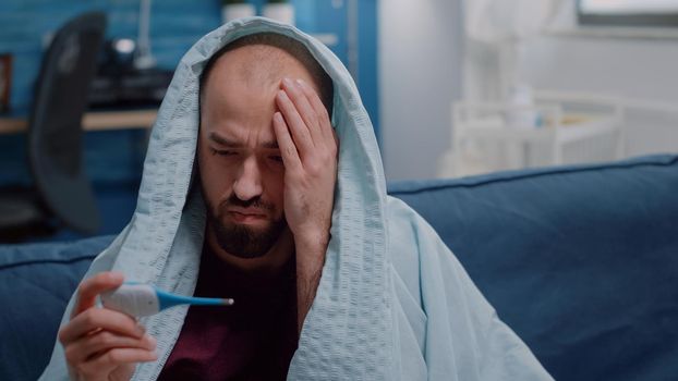 Close up of man wrapped in blanket with thermometer for fever measurement. Sick adult holding device checking temperature to treat virus symptoms and disease. Person with coronavirus