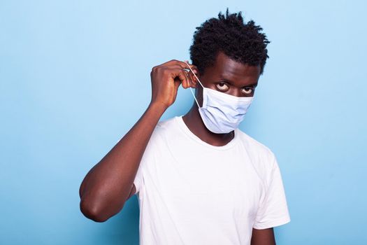 Portrait of person putting mask on face in studio. Casual man looking at camera while using hands to put protective face mask against coronavirus epidemic, standing over blue background