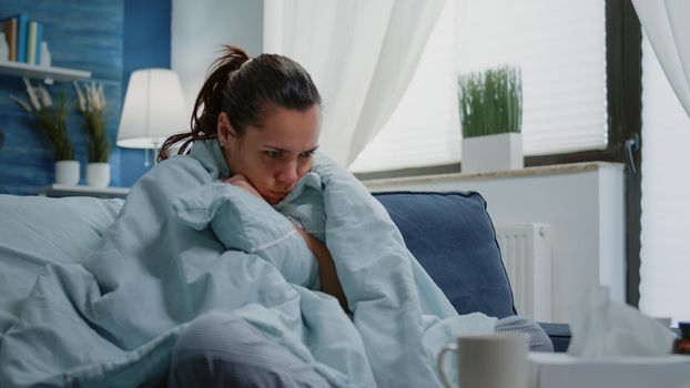 Person with disease feeling cold with blanket and pillow on couch. Sick woman using cover to keep warm having fever and coronavirus symptoms. Adult with illness treatment and medicaments