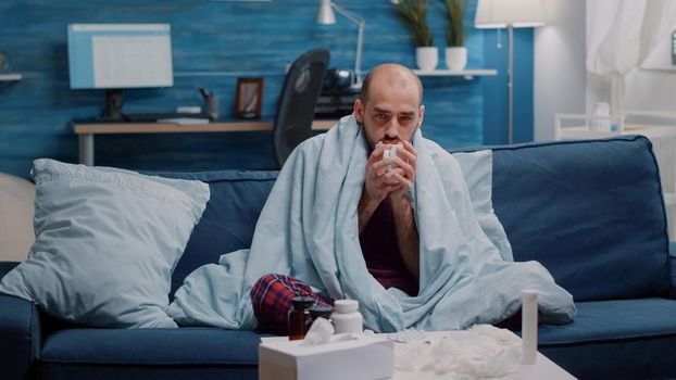 Man with sickness holding cup of tea to cure cold and flu while looking at camera. Portrait of ill person in blanket having medicaments on table and beverage against fever and virus symptoms