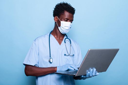 Man nurse with uniform holding laptop over isolated background in studio. Medical assistant with face mask and gloves for coronavirus protection while looking at modern device in hand