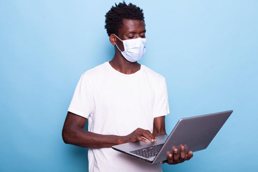 Portrait of casual person looking at laptop and wearing face mask. Man holding modern device while having protection against coronavirus flu pandemic. Young adult with mask and gadget