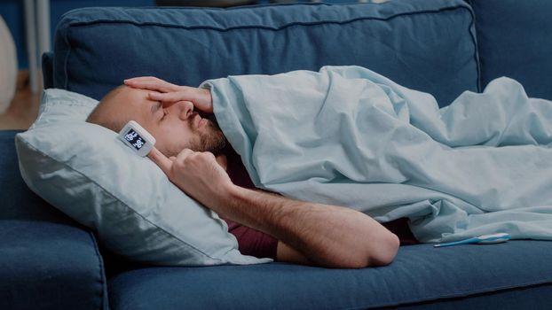 Sick adult using oximeter to measure raising oxygen saturation and pulse pressure. Ill person resting on sofa checking symptoms to treat sickness. Man with disease laying with blanket