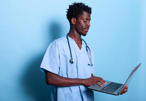 African american man nurse looking at laptop in hand. Black medical assistant with blue uniform and stethoscope holding modern device, standing over isolated background. Healthcare specialist