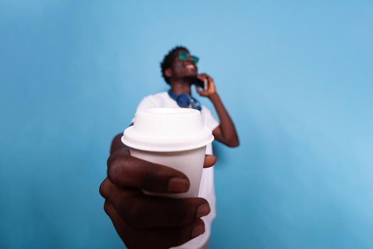 Happy man showing cup of coffee to camera. Casual adult wearing sunglasses and headphones, holding drink while chatting on phone call and feeling carefree. Person having fun with beverage
