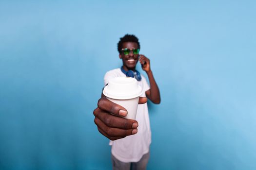 Carefree man holding coffee drink close to camera in studio. Casual adult talking on smartphone call and wearing sunglasses while showing beverage over isolated background. Person smiling