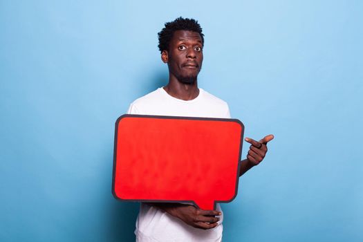 African american man holding red speech bubble in studio. Black person with blank sign for communication and messages on board looking at camera and standing over isolated background