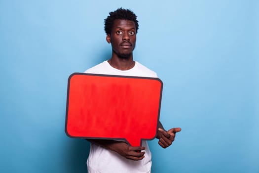 African american man holding red speech bubble in studio. Black adult with blank sign for communication and conversation on board looking at camera and standing over isolated background