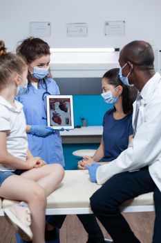 Radiologist nurse holding tablet with heart diagnostic on screen during clinical examination in hospital office. African american pediatrician doctor with protective face mash against coronavirus