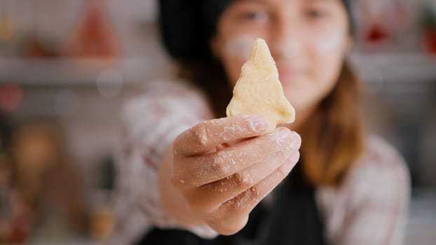 Selective focus of child holding cookie dough with tree shape in hands preparing homemade traditional gingerbread enjoying cooking dessert in culinary kitchen. Girl enjoying christmas holiday