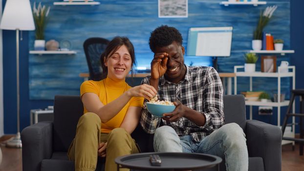 Cheerful interracial couple laughing at comedy on television in living room. Young mixed race partners having fun together, enjoying movie on TV at home. Happy multi ethnic people