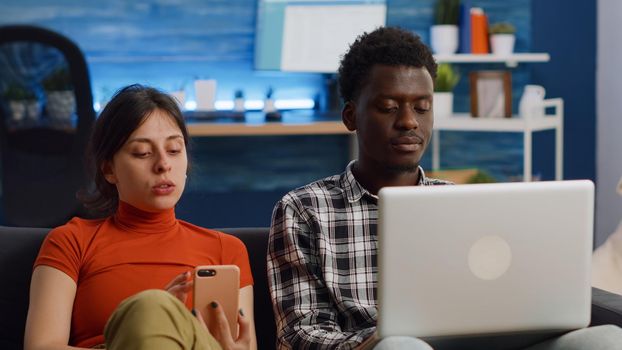 Modern interracial couple using technology on couch at home. Caucasian woman holding smartphone while african american man working on laptop sitting together in living room on sofa