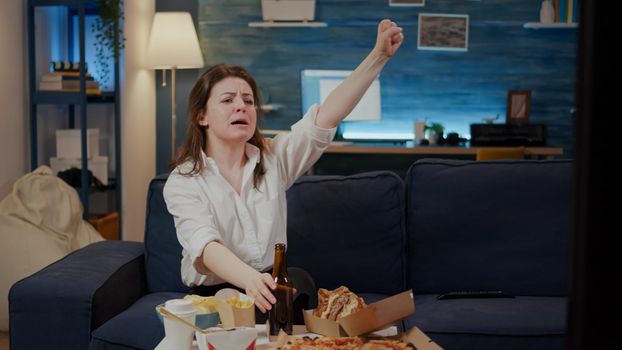 Young adult cheering at football game while eating food from delivery in living room. Caucasian woman watching soccer and enjoying fast food meal with hamburger and bottle of beer