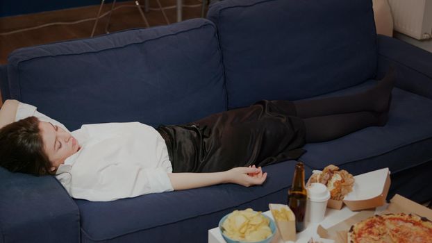 Young person falling asleep on couch dropping TV remote at home. Tired woman from work sleeping after eating takeaway food in living room. Adult taking nap while watching television