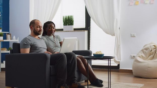 Interracial couple using video call communication at home talking to friends on laptop. African american woman and caucasian man chatting on remote online conference sitting in living room