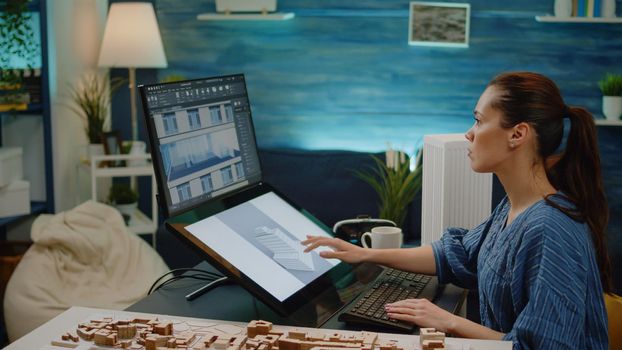 Young engineer using technology for construction design in office. Woman working as architect with building model on touch screen and maquette. Businesswoman analyzing blueprint plan.