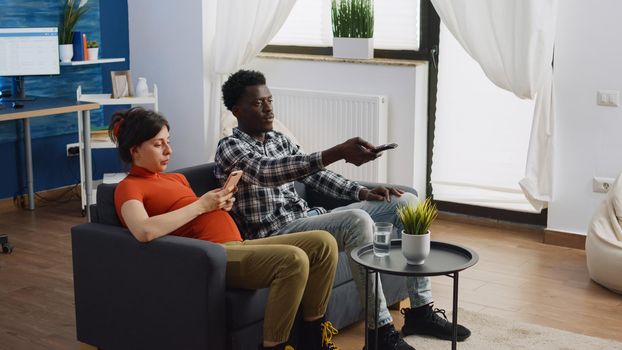 Modern interracial couple with pregnancy using technology in living room. Multi ethnic parents expecting child using smartphone and switching channels on television with remote control