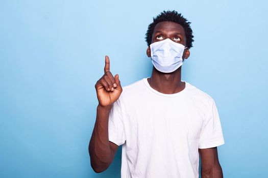 Casual man with face mask against coronavirus pointing up with index finger. Young adult wearing protection for virus pandemic while indicating direction, doing sign with hand.