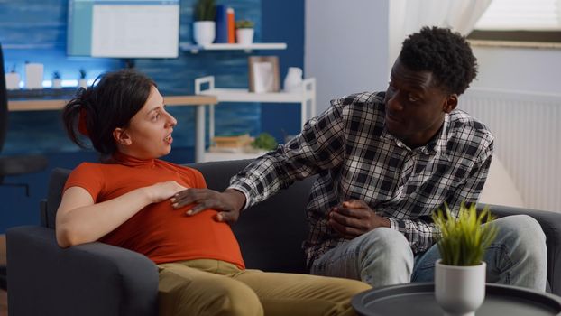 Young interracial couple with pregnancy chatting about baby at home. African american father of child touching belly while pregnant caucasian woman relaxing on sofa in living room.
