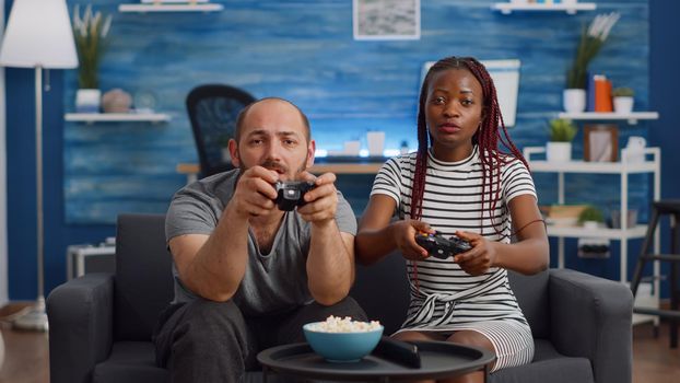 POV of young interracial couple using technology for enjoyment at home. Married multi ethnic man and woman playing video game on TV console with controllers on living room couch