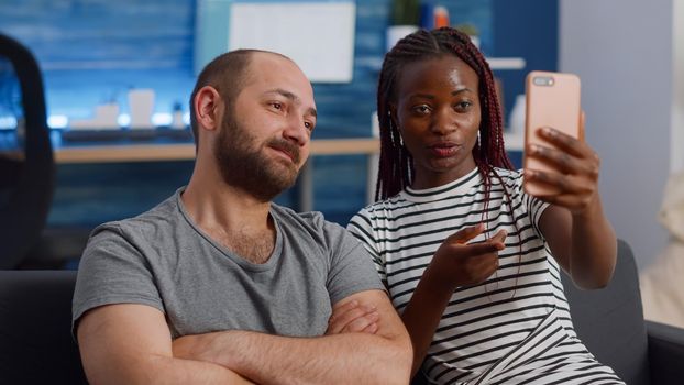 Young interracial couple using online video call conference on smartphone in living room. Modern mixed race lovers talking to friends via internet for remote communication on couch