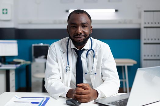 Portrait of african american practitioner doctor working in hospital office checking medical treatment against patient disease. Man physician analyzing medicine prescription using laptop computer