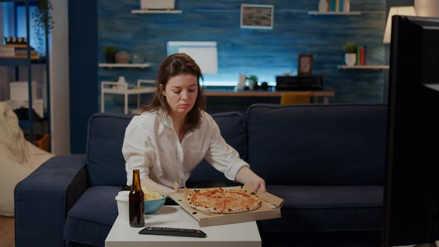 Young adult receiving order of takeaway food sitting at home. Woman with pizza delivered in box feeling hungry while watching television. Person eating fast food as unhealthy dinner