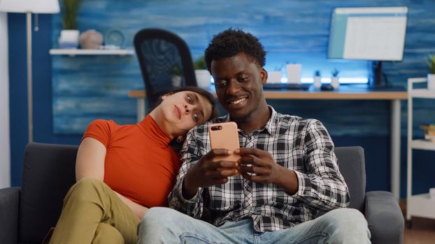 Joyful interracial couple taking selfies with smartphone while sitting together on living room sofa. Cheerful multi ethnic people having fun with pictures and technology at home