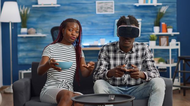 Black couple playing virtual game with vr glasses at home. POV of african american man using headset and joystick for fun activity in living room while woman eating popcorn, watching boyfriend
