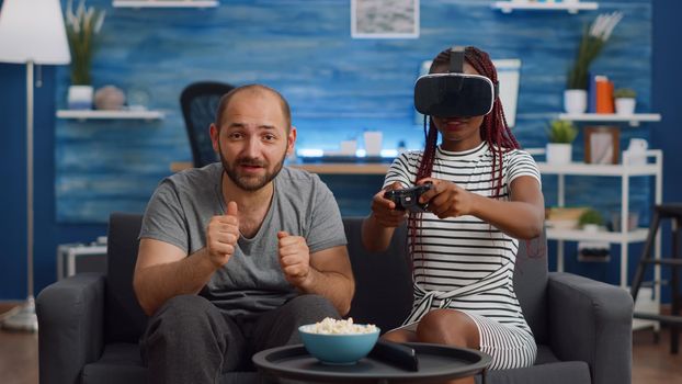 Married interracial couple using technology at home. African american woman with vr glasses playing game while caucasian man watching and cheering in living room. Mixed race cheerful people