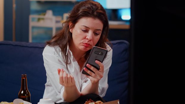 Caucasian woman biting slice of pizza and holding smartphone in living room. Young adult using technology while watching TV and enjoying fast food meal and bottle of beer from delivery