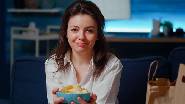Portrait of smiling woman holding bowl of chips in hands while sitting on couch at home. Close up of caucasian person with snack and bag with takeaway food on sofa, looking at camera