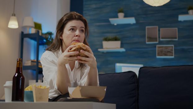 Adult with takeaway order of fast food watching television in living room. Woman eating hamburger and drinking beer from bottle while sitting on couch at home. Person enjoying delivery meal