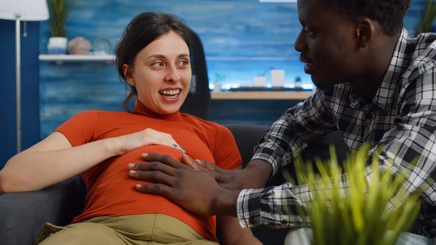 Married interracial couple waiting for baby and parenthood at home. Young multi ethnic man and woman being pregnant with child sitting on couch. African american husband touching belly