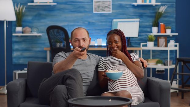 POV of interracial couple with pregnancy watching television and eating popcorn. Multi ethnic people expecting child, sitting on sofa. Partners having fun while looking at camera