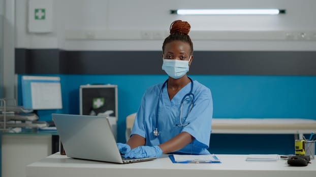 Portrait of medical assistant with uniform and face mask in cabinet at healthcare facility. Young nurse sitting at desk working on laptop in office during coronavirus pandemic