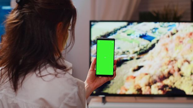 Caucasian person vertically holding green screen on smartphone in living room. Young woman looking at chroma key display with mockup template and isolated background on modern gadget