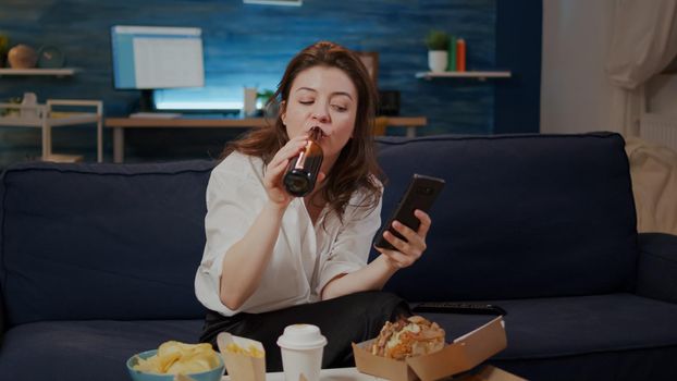 Person eating pizza and looking at smartphone screen sitting on couch. Caucasian woman with internet technology watching TV while enjoying takeaway fast food meal in living room