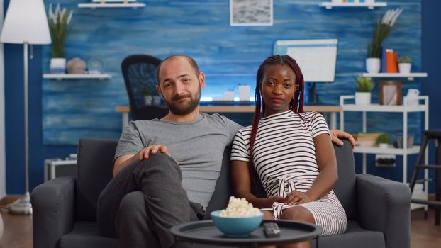Interracial couple waving at video call camera and talking to friends at home. Multi ethnic married people using online conference for remote communication while sitting in living room.