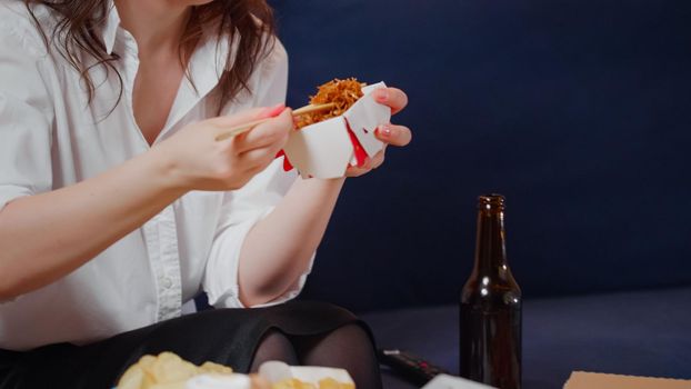 Young woman taking box of chinese food from table to eat using chopsticks while watching movie on television. Person relaxing after work with fast food takeaway in living room at home