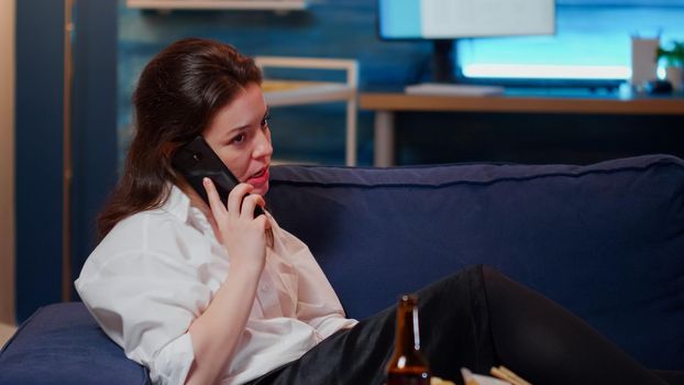 Business woman eating takeaway asian food on sofa while boss calling on smartphone. Annoyed adult trying to enjoy free time when phone ringing at home. Person with chinese meal after work