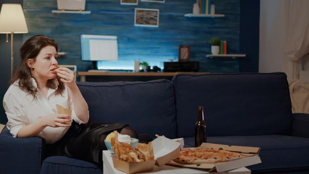 Woman relaxing on couch with takeaway food at home in living room. Young person having big fast food meal and laying on sofa watching TV while eating fries from delivery package