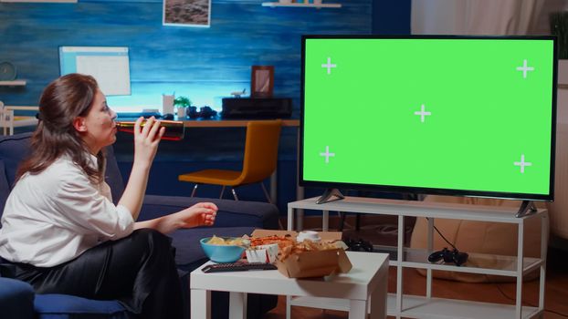 Caucasian woman watching green screen on television while eating chips and drinking beer. Person sitting on couch watching horizontal mockup template and isolated background while laughing