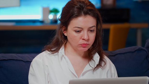 Close up of woman eating chinese food with chopsticks while working on laptop in living room at home. Young adult using technology for business while watching TV and enjoying takeaway meal