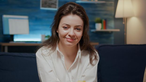 Caucasian adult looking at camera while having bowl of chips in hand sitting on couch in living room. Portrait of young woman with delivery bags and package on sofa and table at home