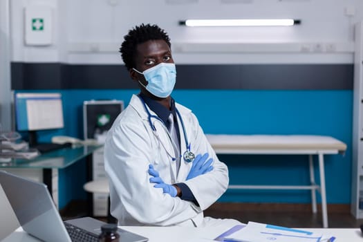 Doctor of african american ethnicity sitting at desk with face mask and stethoscope looking at camera in medical cabinet. Black medic wearing gloves and coat at healthcare clinic