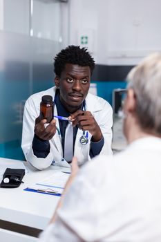 African american doctor showing jar of healing pills to elderly woman sitting in medical cabinet. Old patient receiving treatment, medicine, drugs from healthcare specialist at desk